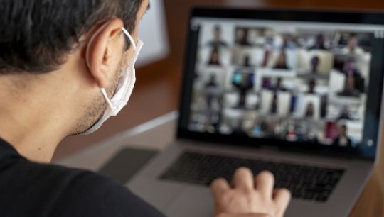 Guy with a mask on while looking at his laptop that has a zoom meeting up with tens of people on it