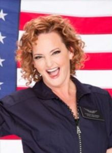 Woman with curly red hair and a navy blue jacket smiling brightly with an American flag behind her | Elizabeth McCormick